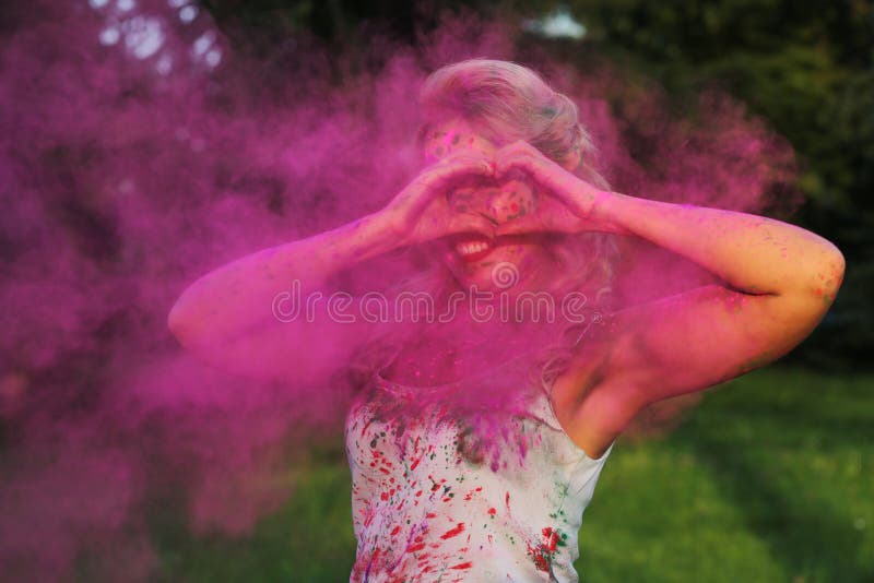 Smiling blonde model having fun in a cloud of pink dry Holi paint and showing heart sign from her fingers. Smiling blonde woman having fun in a cloud of pink dry stock images